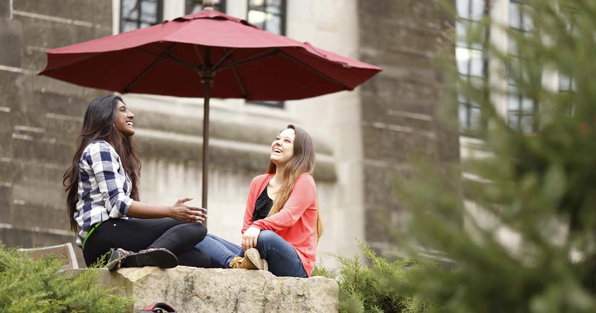 Two students sit and laugh together in front of a campus building. 