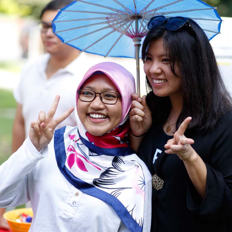 Two students make peace signs while holding an umbrella outside. 