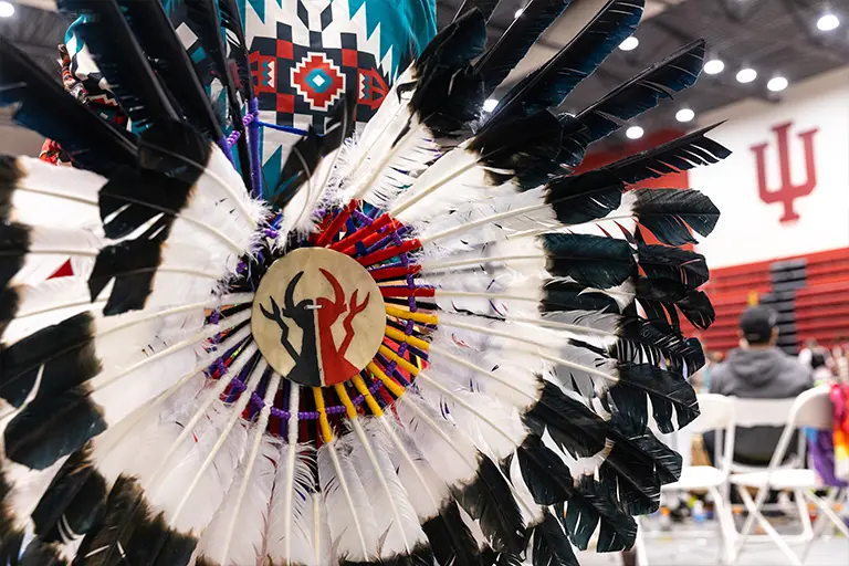 A close-up shot of a traditional Native American headdress with white and black feathers, adorned with a circular medallion featuring a black silhouette of two figures against a beige background, with red and yellow accents. The headdress is displayed against a backdrop of a blurred gymnasium interior with red seating